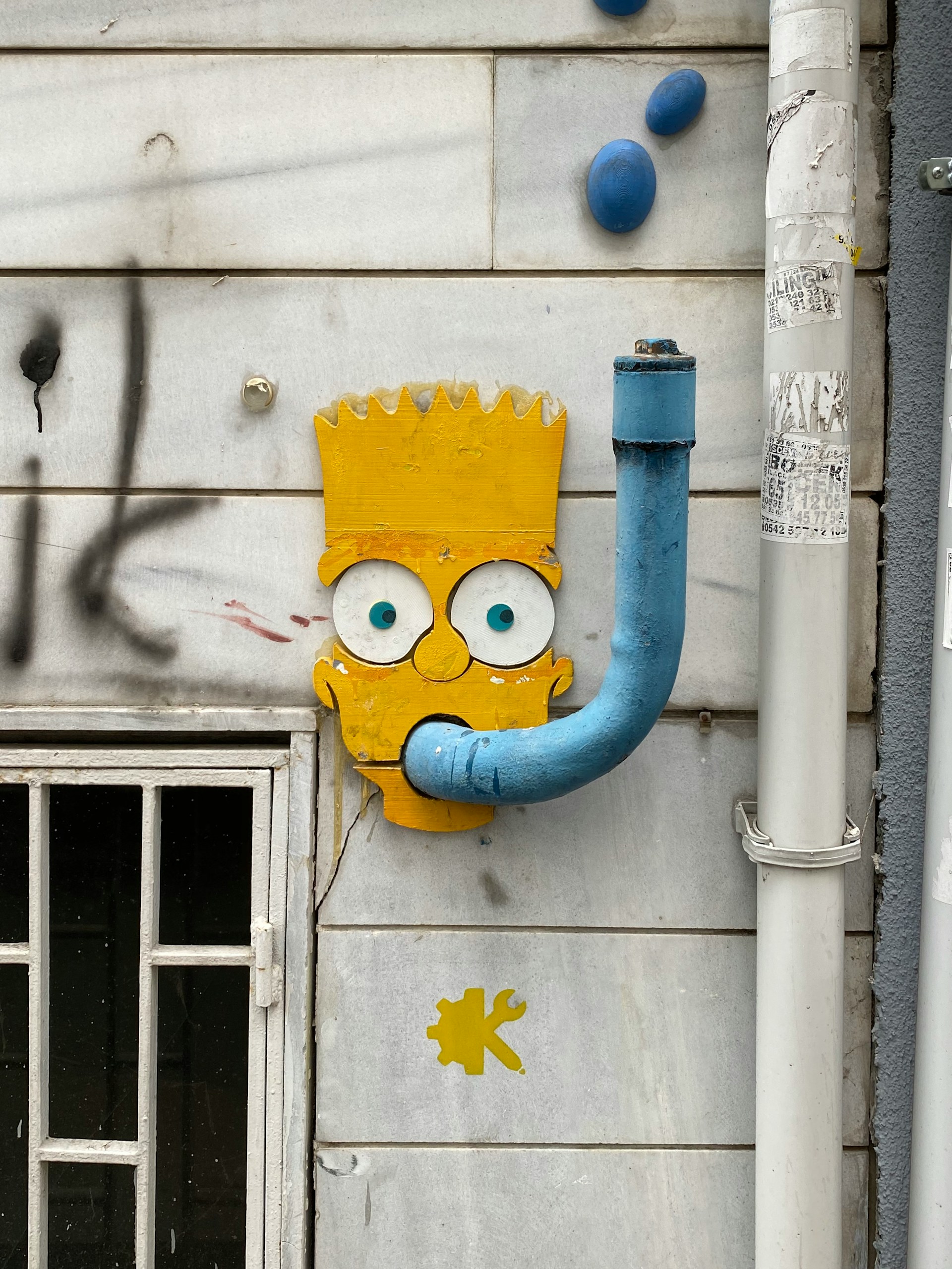 Playful street-art face made from a pipe and painted wood on a city wall
