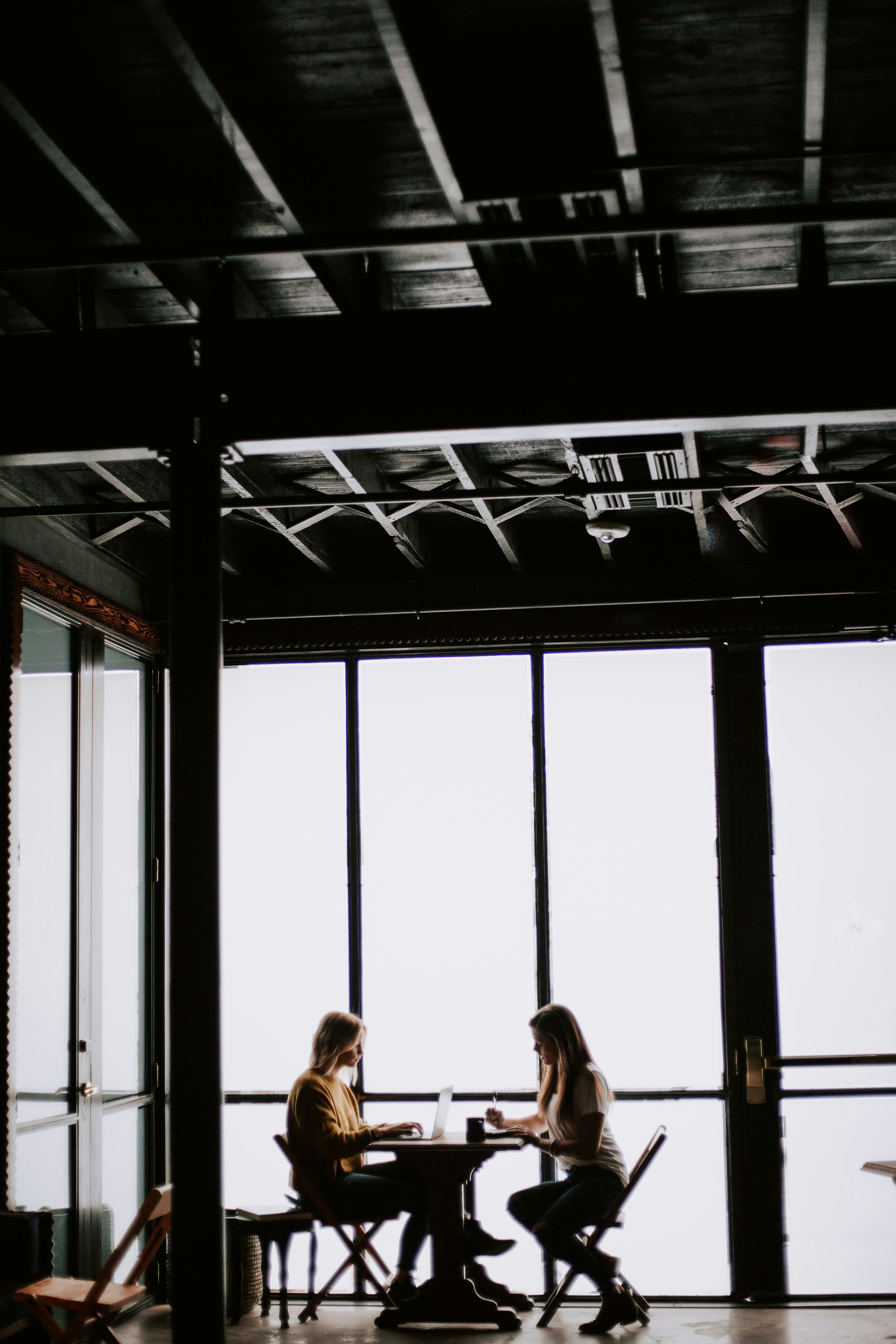 Two people in conversation at a table beside tall industrial windows